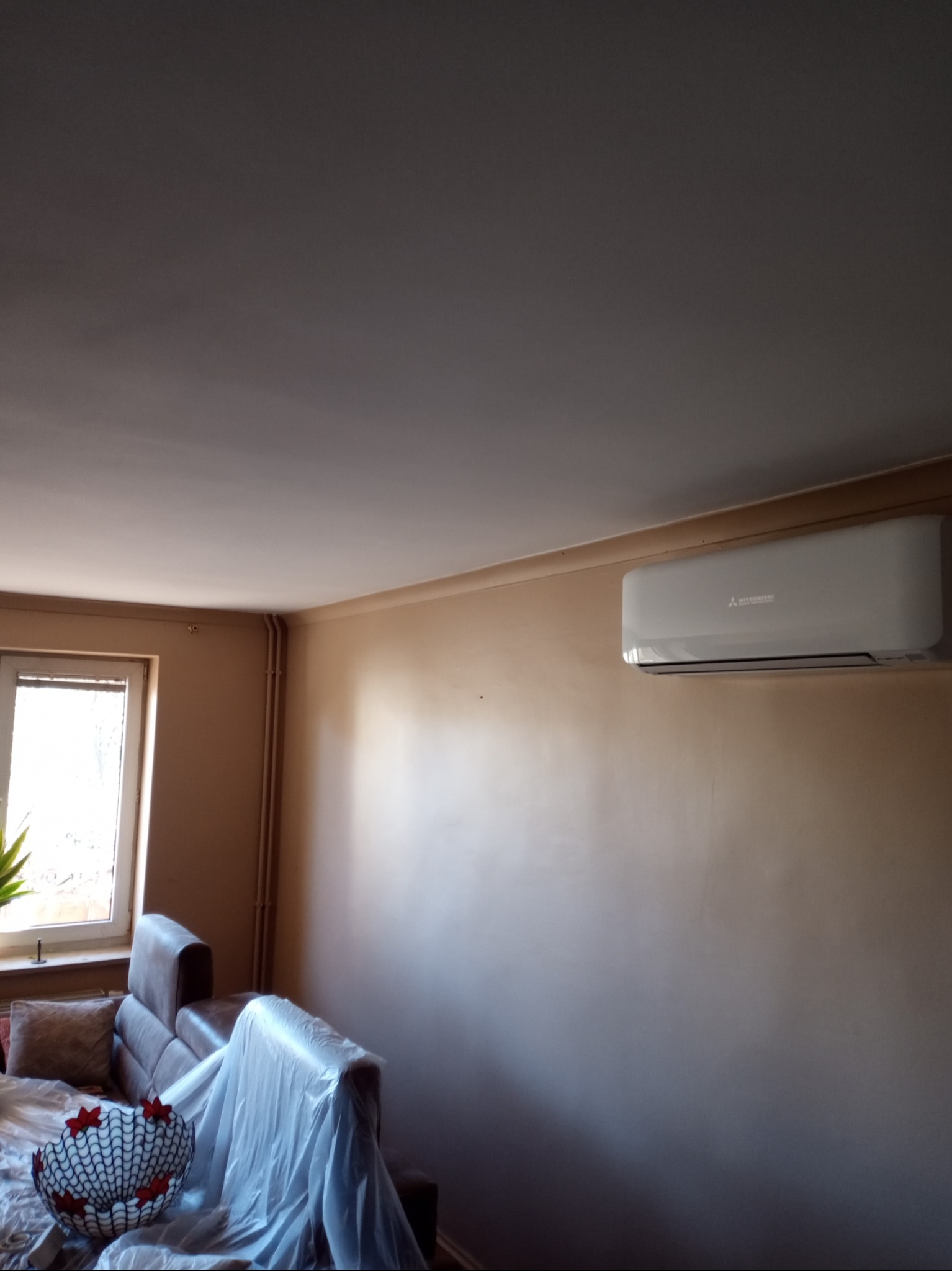 Beżowa ściana w pokoju z zamontowanym klimatyzatorem Mitsubishi, zabezpieczone meble folią malarską.