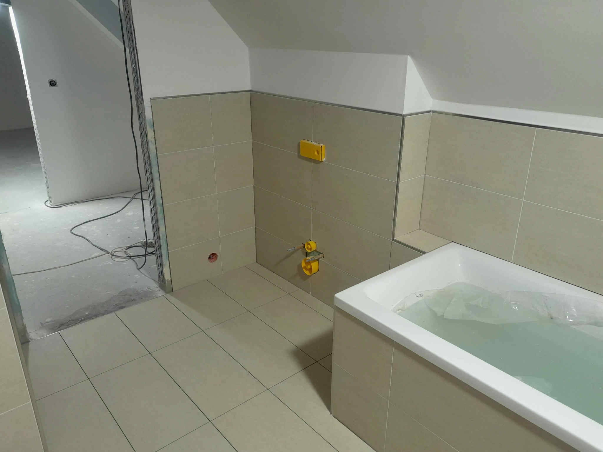 Beżowe płytki ścienne i podłogowe w łazience na poddaszu, z białą wanną wypełnioną wodą. Żółte elementy instalacji wodnej na ścianie. Widok na surowy stan drugiego pomieszczenia.