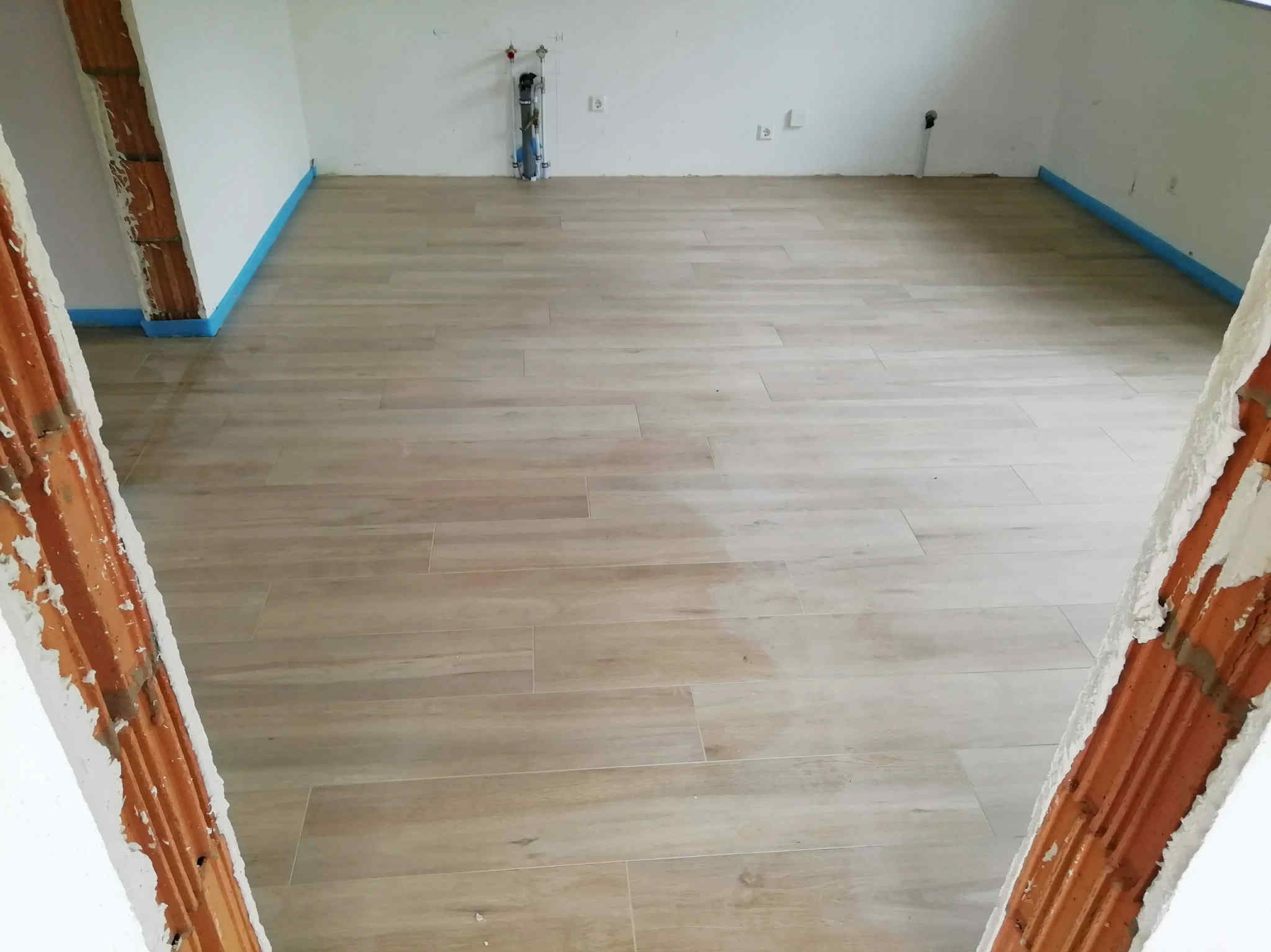 Jasne, drewnopodobne panele ułożone w pustym pomieszczeniu z fragmentami surowych ścian. Niebieska taśma zabezpieczająca przy podłodze. Widoczne instalacje na ścianie.