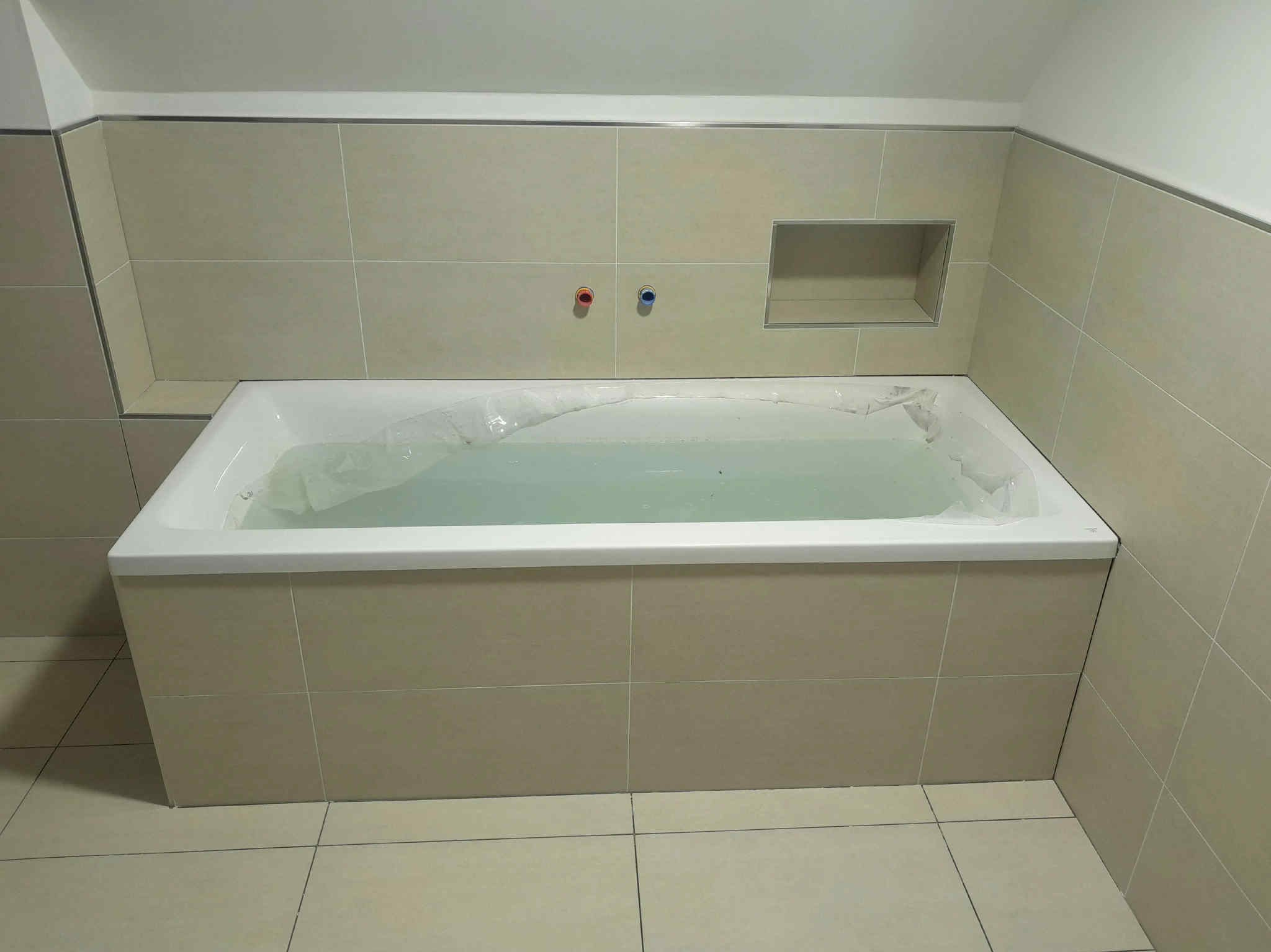 Wykończona łazienka z wanną obłożoną beżowymi płytkami, widoczne baterie i wnęka w ścianie. Minimalistyczny design, jasne wnętrze.