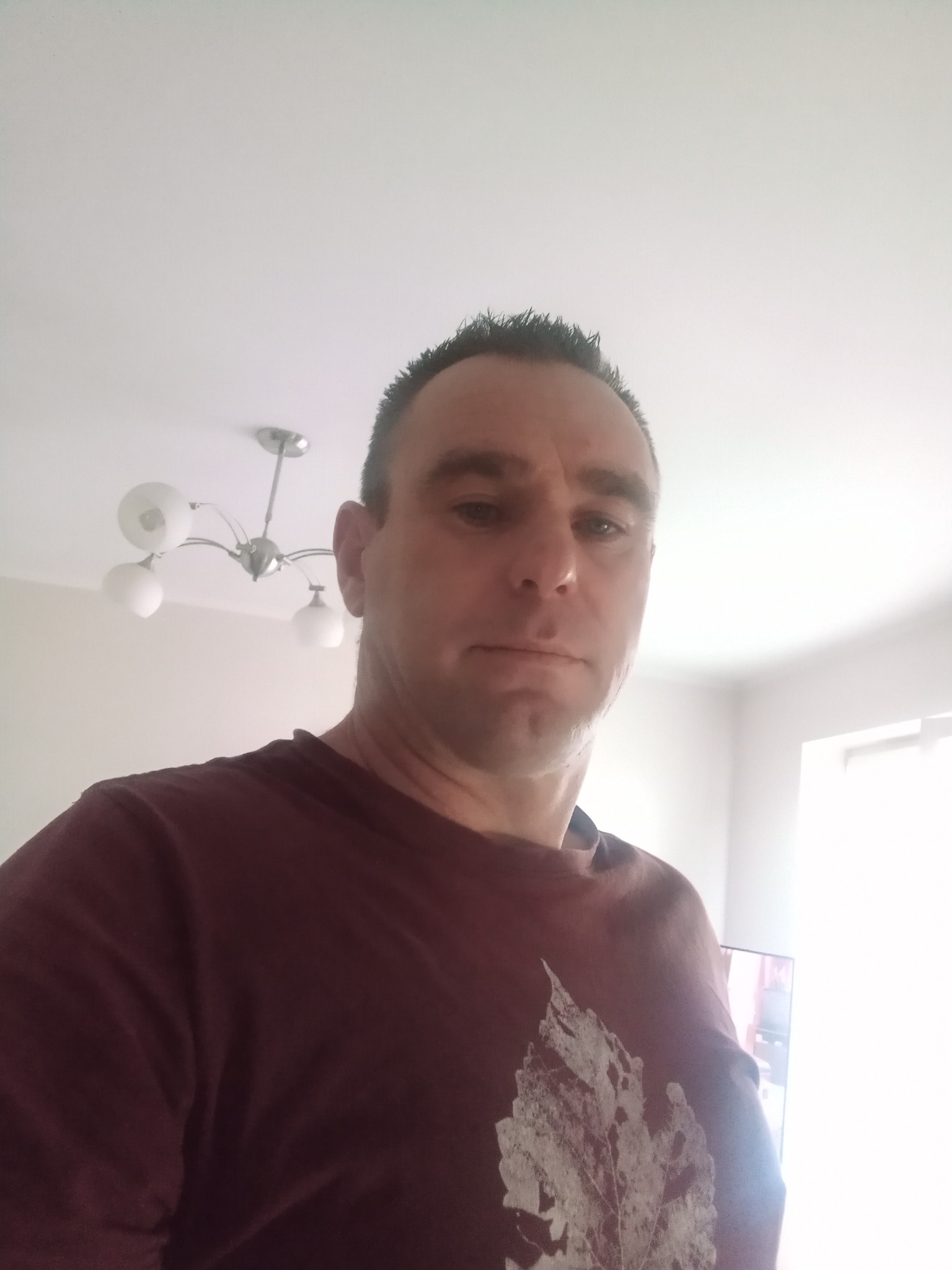 Mężczyzna w bordowej koszulce z nadrukiem liścia, patrzący w obiektyw, z widoczną lampą sufitową i fragmentem okna w tle. Ujęcie z dołu.