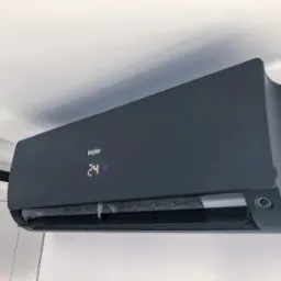 Czarny klimatyzator Haier zamontowany na białej ścianie, wyświetlacz pokazuje 24 stopnie Celsjusza, widoczne żaluzje w tle.