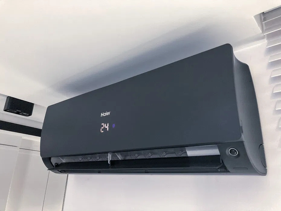Czarny klimatyzator Haier zamontowany na białej ścianie, wyświetlacz pokazuje 24 stopnie Celsjusza, widoczne żaluzje w tle.
