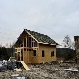 VALERII ROMANIUK - Budowa domu w stanie surowym otwartym, widoczna konstrukcja dachu z deskowaniem, robotnik na dachu, materiały budowlane na ziemi, teren budowy ogrodzony siatką.