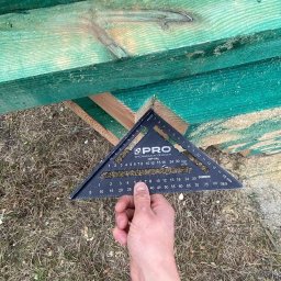 VALERII ROMANIUK - Pomiar kąta desek konstrukcji dachowej za pomocą kątownika stolarskiego PRO, widok z dołu na zielone deski i dłoń trzymającą narzędzie.