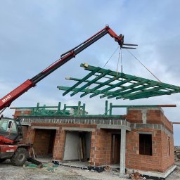 VALERII ROMANIUK - Czerwony dźwig MANITOU podnosi zieloną, drewnianą konstrukcję więźby dachowej nad budynkiem z czerwonej cegły w trakcie budowy.