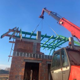 VALERII ROMANIUK - Czerwony dźwig podnosi betonowy element konstrukcyjny na szczyt murowanej ściany budynku z częściowo ułożoną, zieloną więźbą dachową na tle błękitnego nieba.