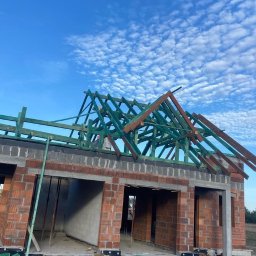 VALERII ROMANIUK - Wznoszenie więźby dachowej na murowanym domu w stanie surowym otwartym, widoczne elementy drewniane pomalowane na zielono, błękitne niebo w tle.