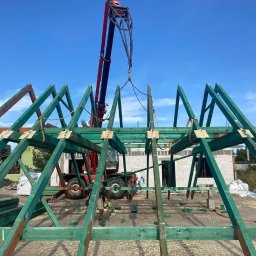 VALERII ROMANIUK - Montaż drewnianej konstrukcji dachu z użyciem dźwigu Manitou, widoczne elementy więźby dachowej pomalowane na zielono, w tle budynek w trakcie budowy z białego bloczka.