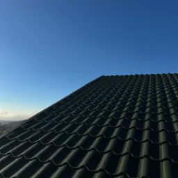 Nowy, ciemny dach z blachodachówki, częściowo pokryty drobnymi kroplami wody, na tle błękitnego nieba.