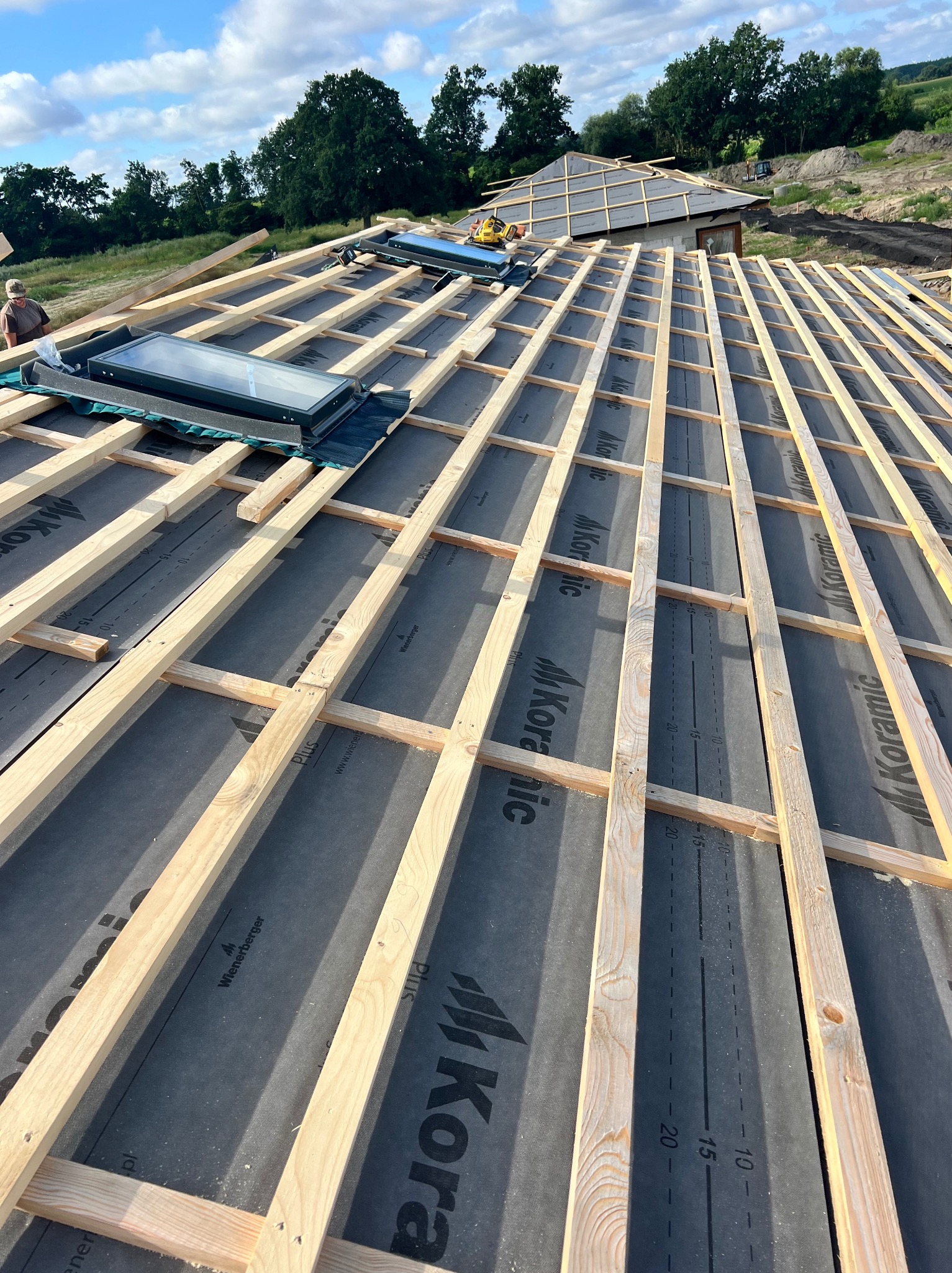 Konstrukcja drewniana pod dach z zamontowanymi oknami dachowymi, widoczna czarna membrana z logo producenta, w tle zalesiony teren.