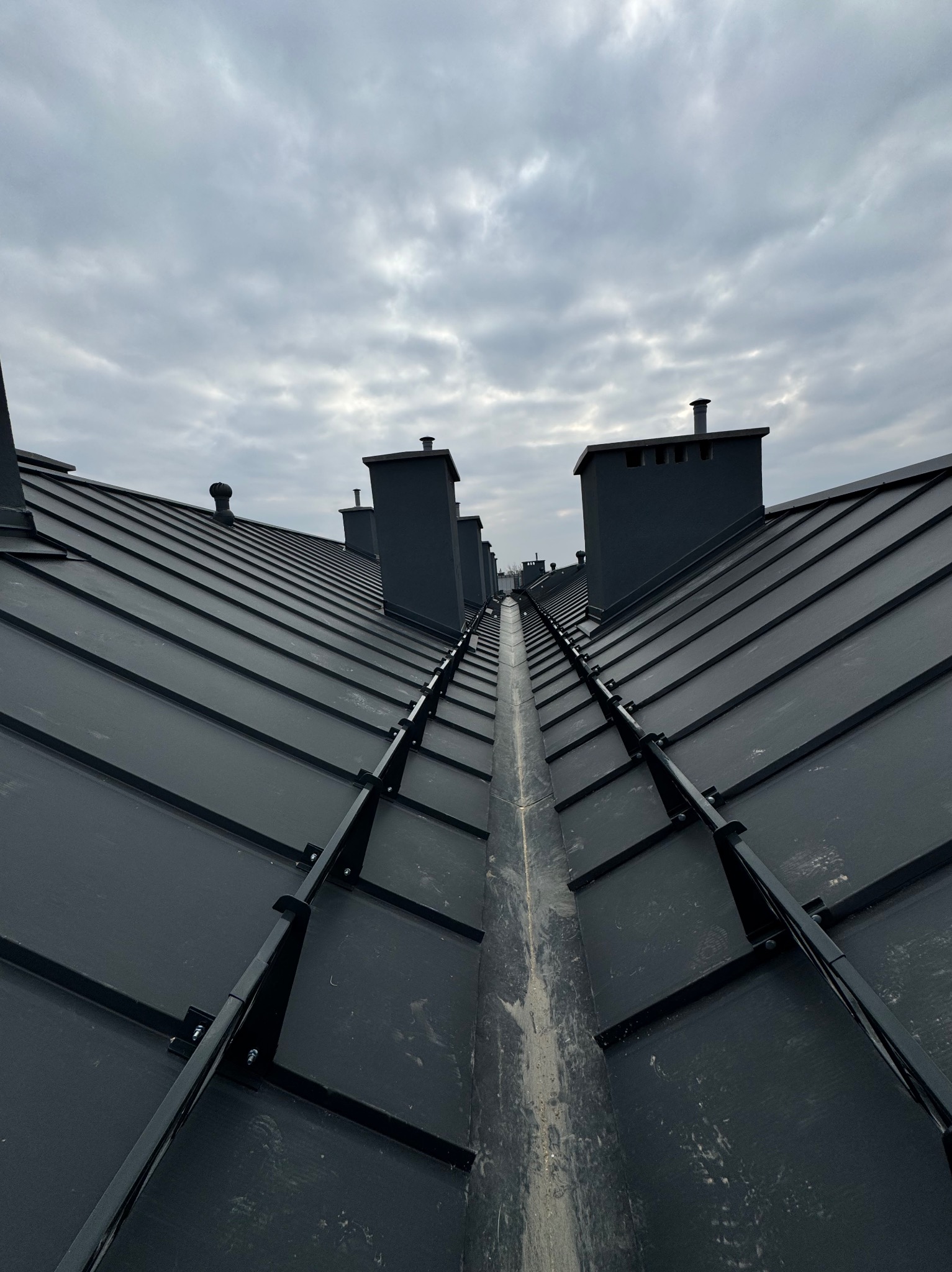 Nowe, czarne, blaszane pokrycie dachowe z widocznymi kominami i rynną w centralnej części kadru, uchwycone z perspektywy żabiej w pochmurny dzień.