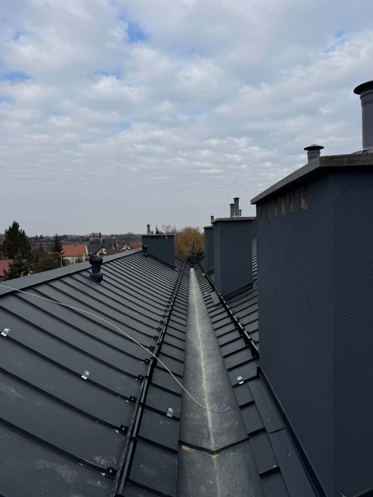 Widok z góry na nowy, grafitowy dach z blachy na rąbek stojący, z widocznymi kominami i elementami mocującymi, na tle pochmurnego nieba i zabudowań miejskich.