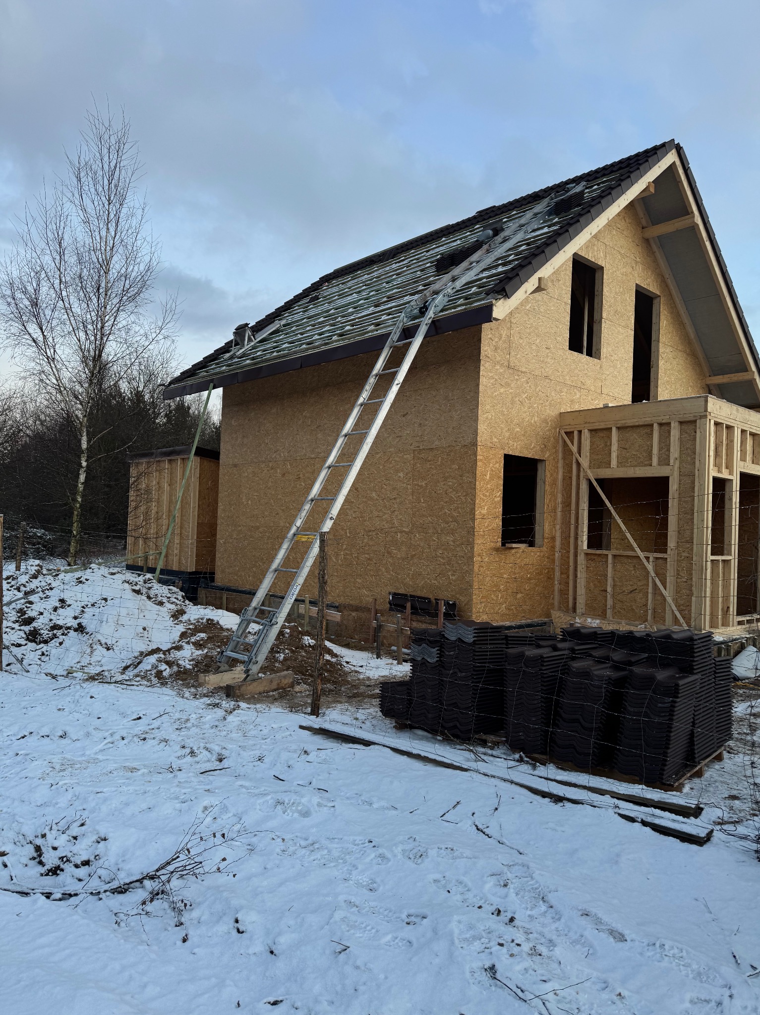 Budowa domu w zimowej scenerii: widoczna konstrukcja z płyt OSB, częściowo pokryty dach z ułożonymi dachówkami, drabina oparta o budynek, stos dachówek na palecie w śniegu.