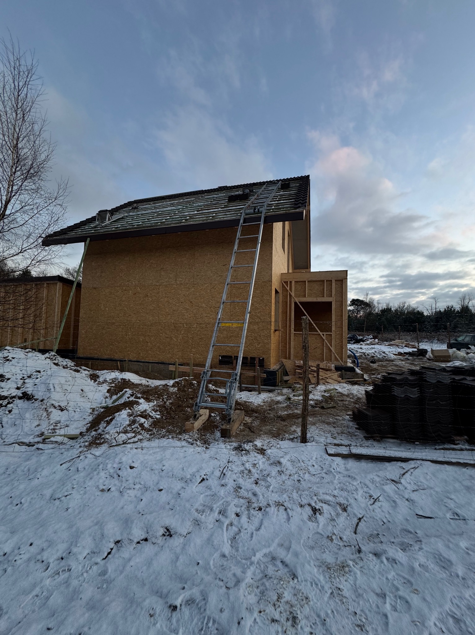 Dom w trakcie budowy w zimowej scenerii, widoczna konstrukcja dachu z łatami i częściowo położonym pokryciem, drabina oparta o ścianę.