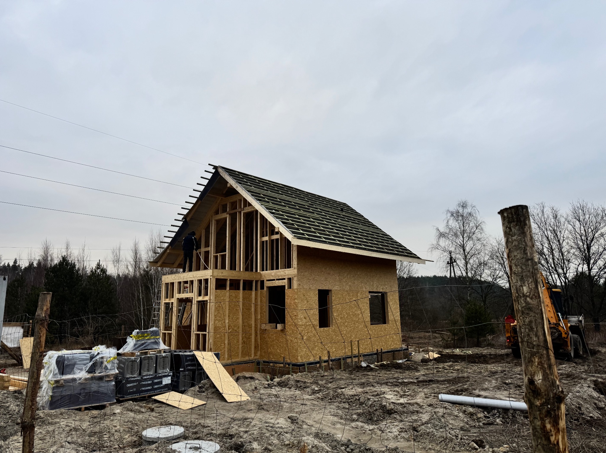 Budowa domu w stanie surowym otwartym, widoczna konstrukcja dachu z deskowaniem, robotnik na dachu, materiały budowlane na ziemi, teren budowy ogrodzony siatką.