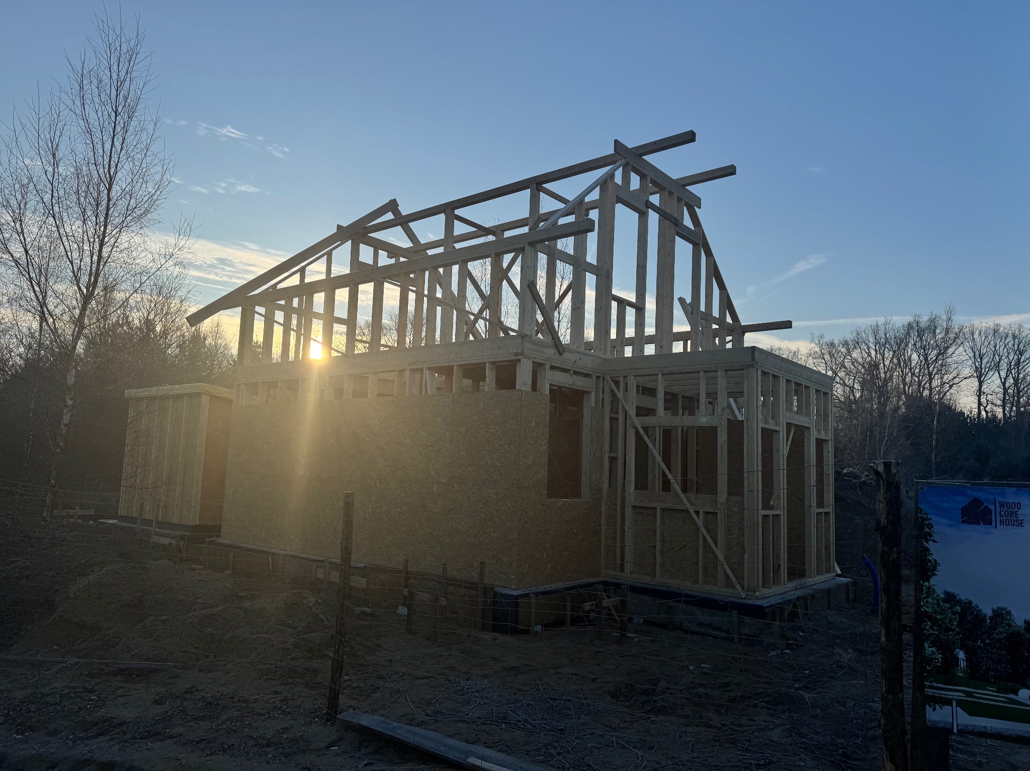 Konstrukcja drewniana domu jednorodzinnego w trakcie budowy, widoczne elementy więźby dachowej i ścian szkieletowych, w tle zachodzące słońce.