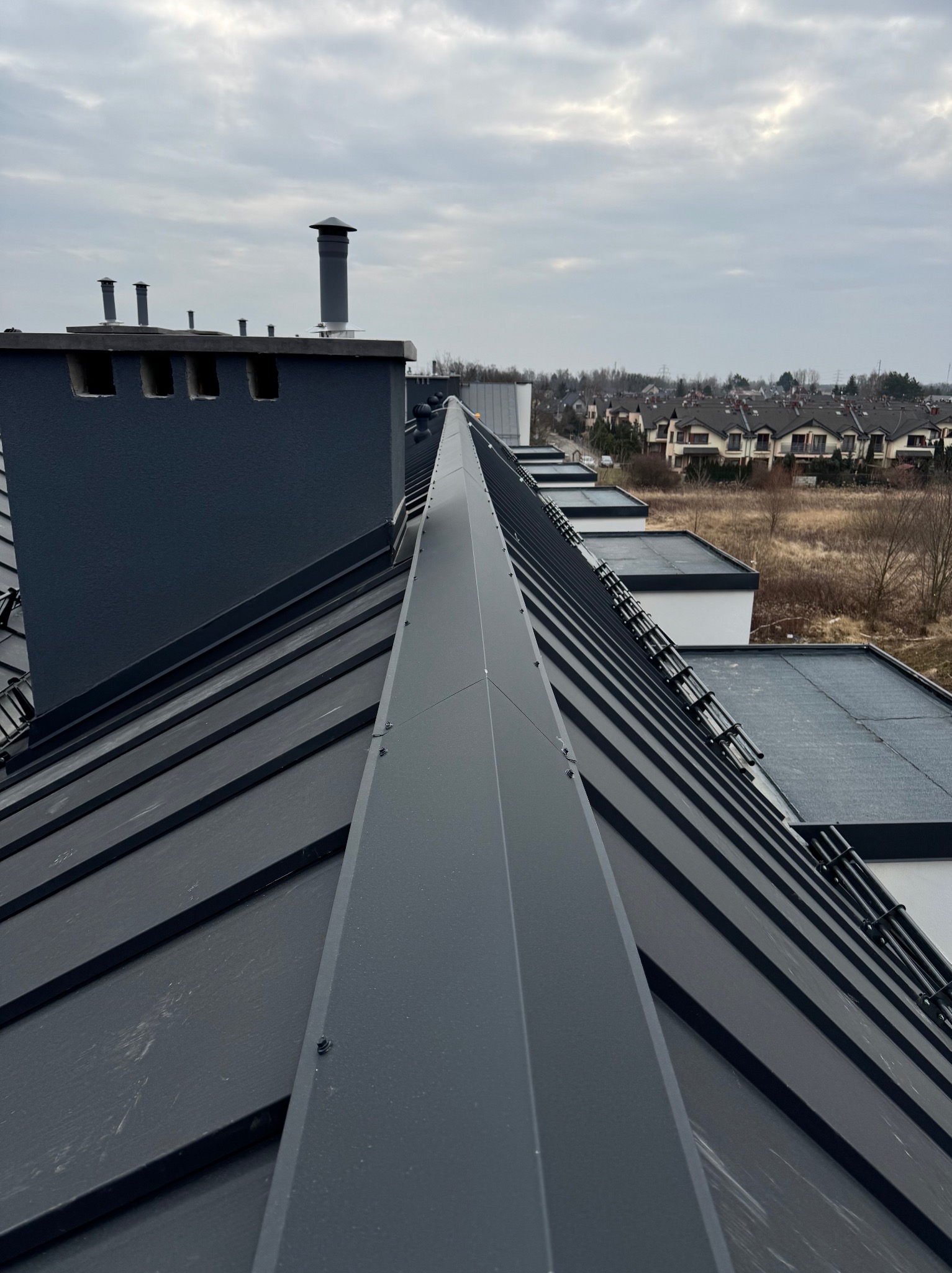 Nowoczesny, grafitowy dach z blachy na tle osiedla domów jednorodzinnych i pochmurnego nieba, widoczne kominy i obróbki blacharskie.