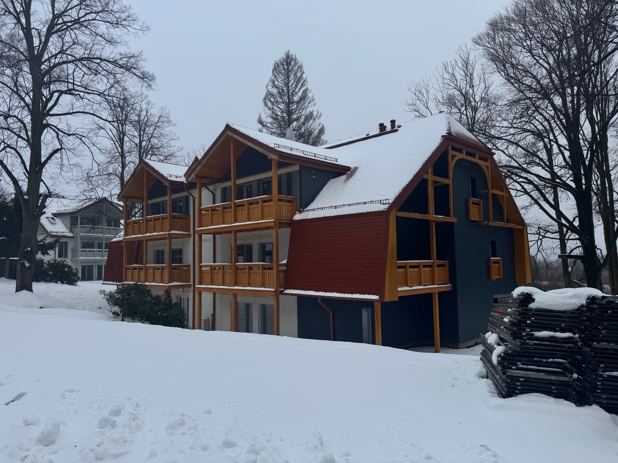 Nowo wybudowany, wielopiętrowy budynek z drewnianymi balkonami i czerwonym dachem pokrytym śniegiem w zimowej scenerii. Obok budynku sterta rusztowań.