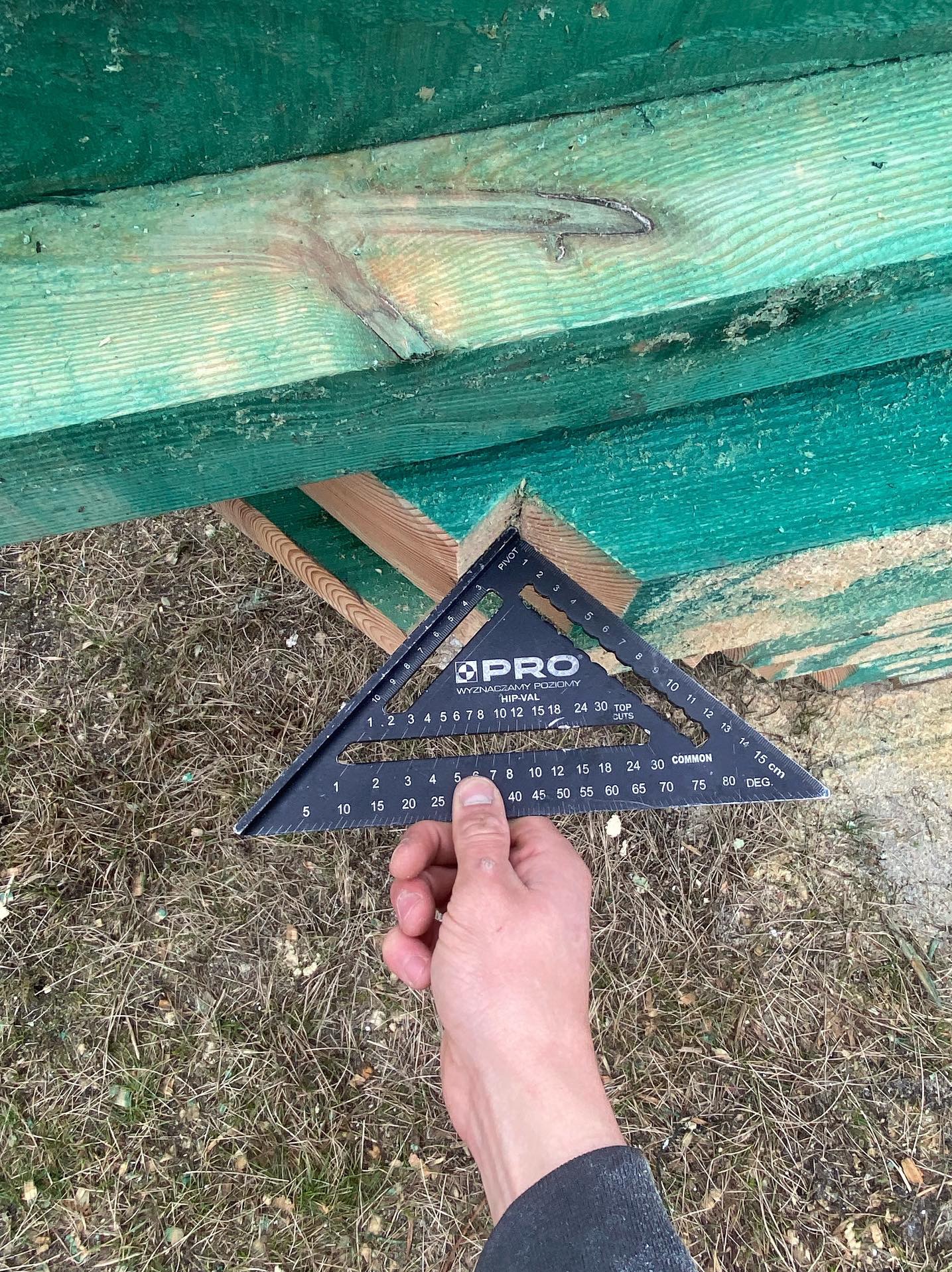 Pomiar kąta desek konstrukcji dachowej za pomocą kątownika stolarskiego PRO, widok z dołu na zielone deski i dłoń trzymającą narzędzie.