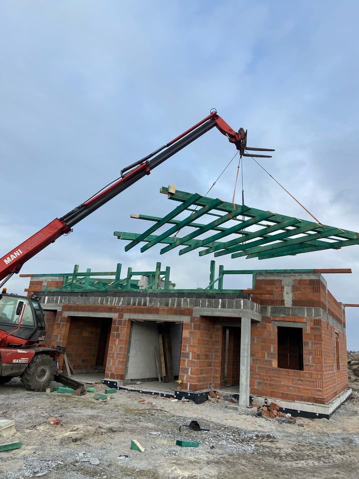 Czerwony dźwig MANITOU podnosi zieloną, drewnianą konstrukcję więźby dachowej nad budynkiem z czerwonej cegły w trakcie budowy.