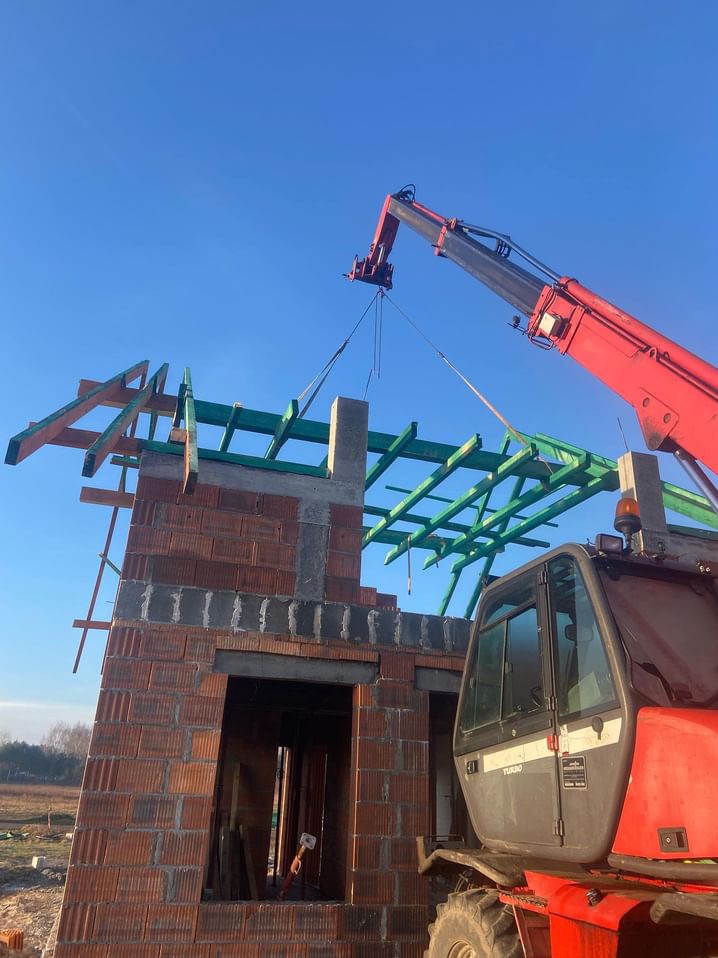 Czerwony dźwig podnosi betonowy element konstrukcyjny na szczyt murowanej ściany budynku z częściowo ułożoną, zieloną więźbą dachową na tle błękitnego nieba.