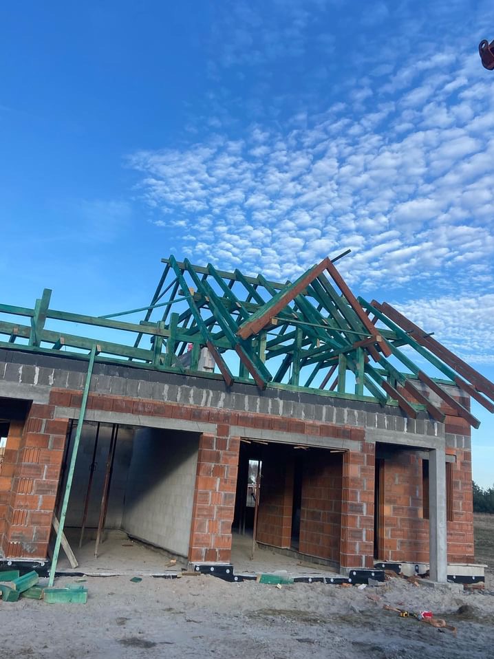 Wznoszenie więźby dachowej na murowanym domu w stanie surowym otwartym, widoczne elementy drewniane pomalowane na zielono, błękitne niebo w tle.