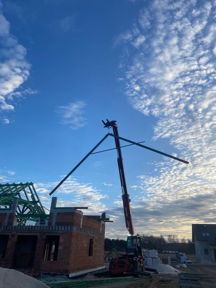 Montaż elementów konstrukcyjnych dachu za pomocą dźwigu na placu budowy domu jednorodzinnego, widoczne niebo z chmurami w tle.