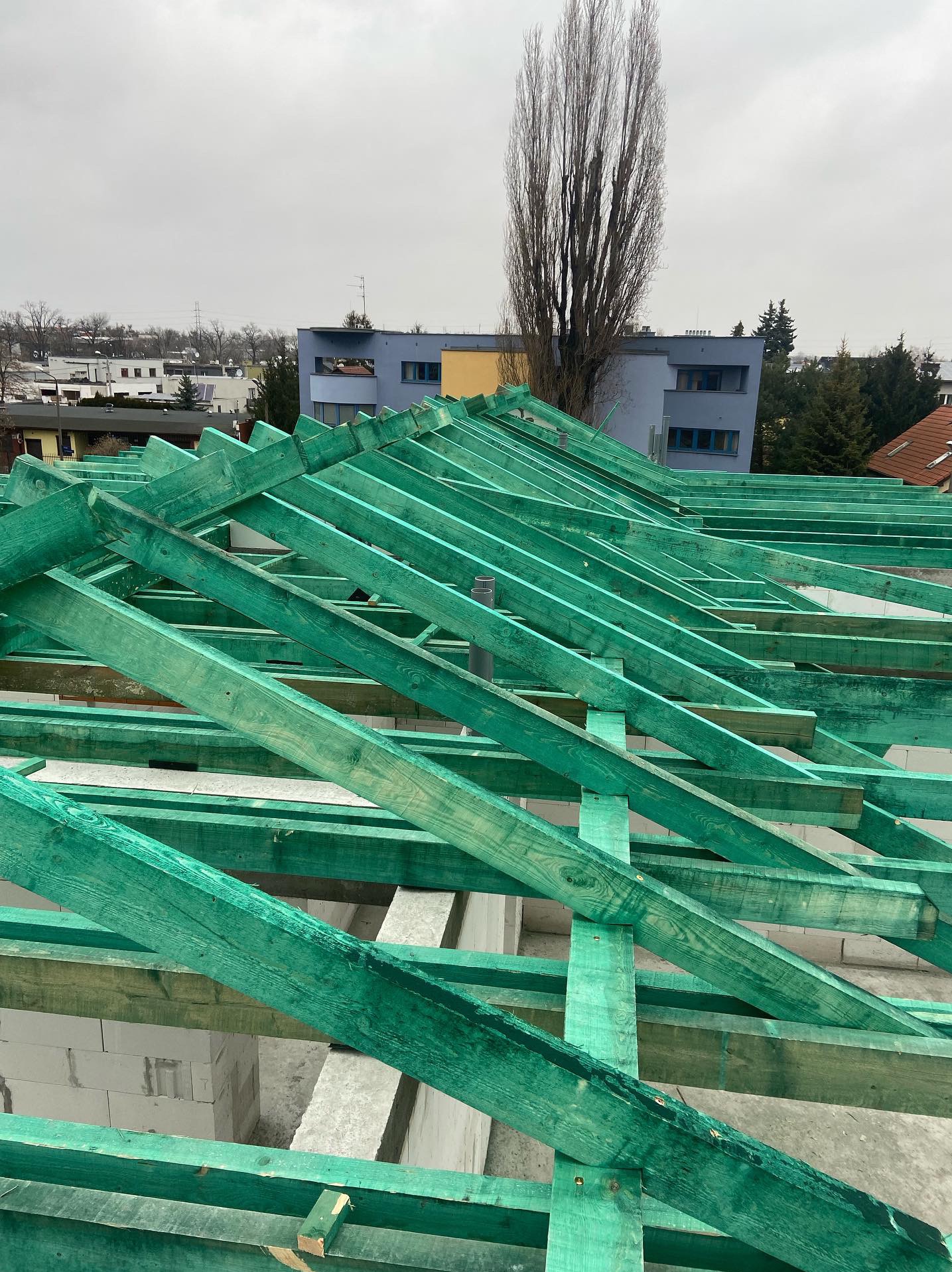 Konstrukcja dachu z impregnowanego na zielono drewna na budynku z betonu komórkowego, widok z góry, w tle zabudowa miejska i pochmurne niebo.