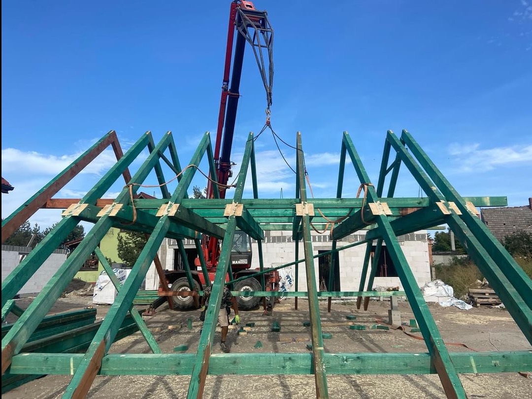 Montaż drewnianej konstrukcji dachu z użyciem dźwigu Manitou, widoczne elementy więźby dachowej pomalowane na zielono, w tle budynek w trakcie budowy z białego bloczka.