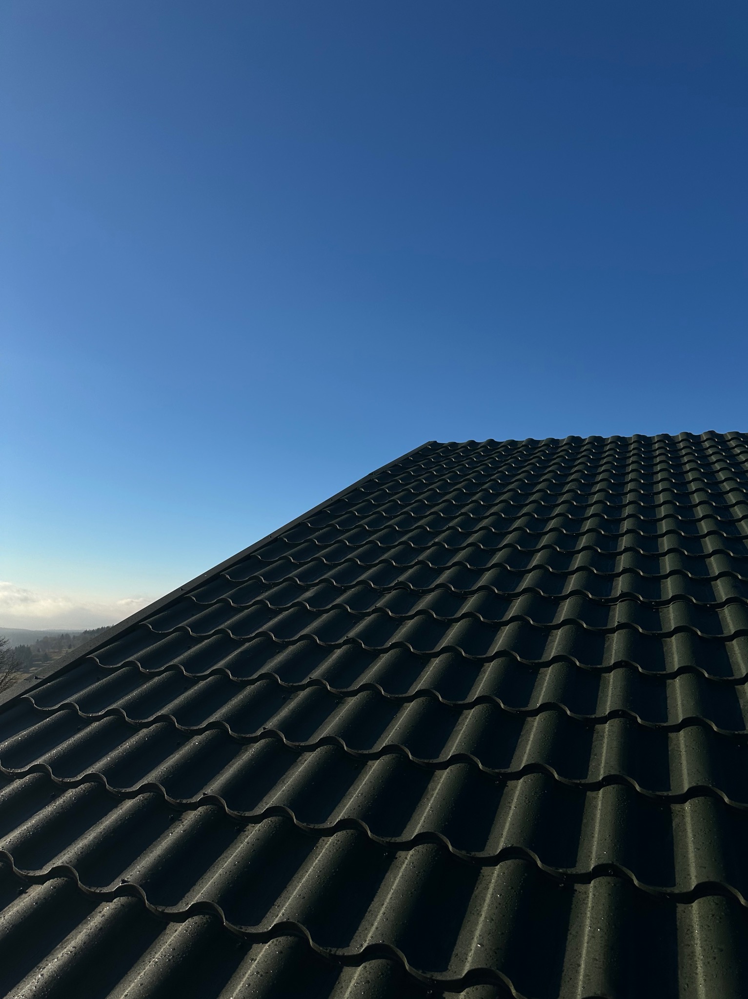 Nowy, ciemny dach z blachodachówki, częściowo pokryty drobnymi kroplami wody, na tle błękitnego nieba.