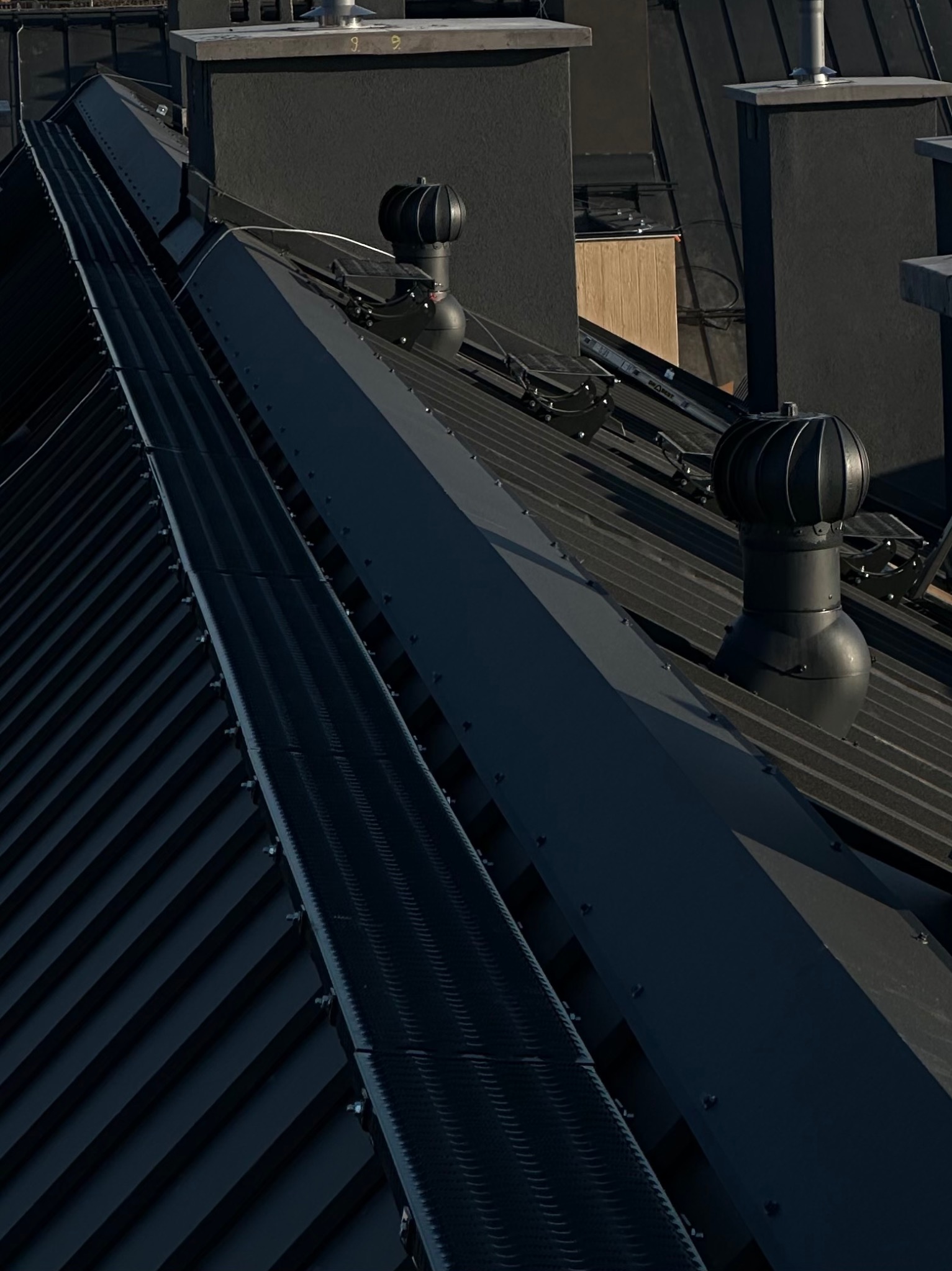 Ciemny, metalowy dach z kominami i turbinami wentylacyjnymi, widoczny z góry pod kątem, z wyraźnymi liniami blachy i elementami mocującymi.