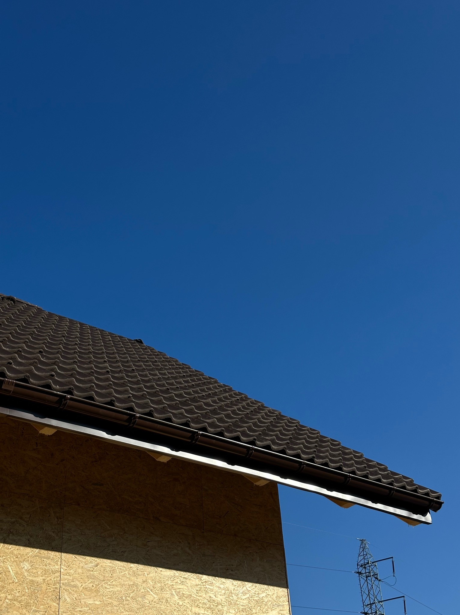 Nowy dach z brązowej blachodachówki trapezowej, widoczna rynna, ściana budynku z płyt OSB i słup wysokiego napięcia na tle błękitnego nieba.