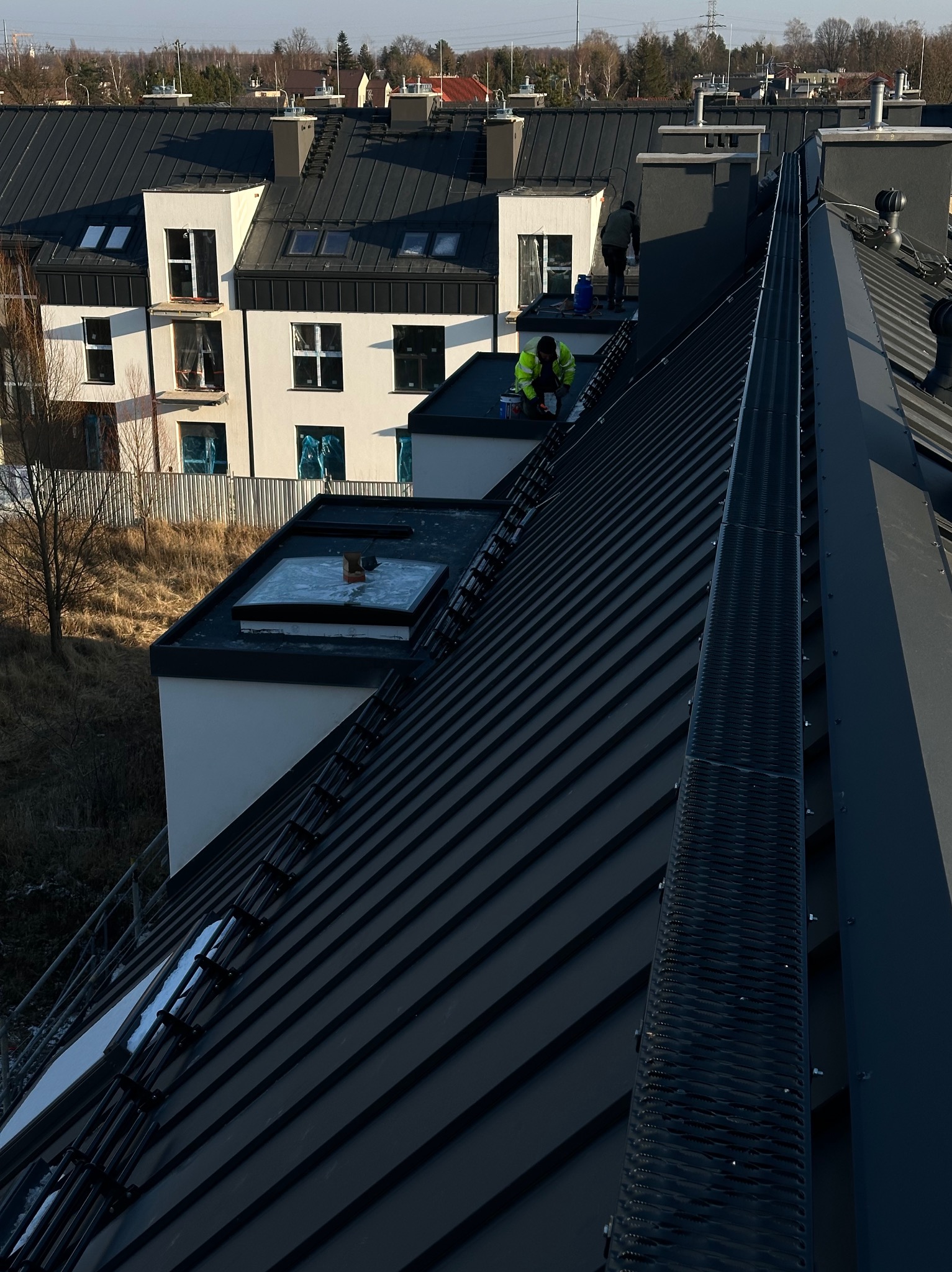 Prace na dachu z blachy na rąbek stojący, widoczni dwaj dekarze w trakcie pracy, jeden w kamizelce odblaskowej, w tle zabudowa szeregowa.