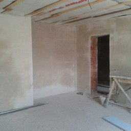 DnD - Pokój w trakcie wygładzania ścian, widoczne surowe wykończenie, drewniane belki sufitowe z folią, ściany pokryte gładzią, drewniany kozioł malarski i fragment niedokończonego przejścia z ceglaną...