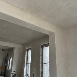 DnD - Wnętrze w trakcie prac wykończeniowych, ściany i sufit pokryte świeżo nałożoną gładzią, widoczne okna w trakcie zabezpieczania folią, na podłodze narzędzia i materiały budowlane.