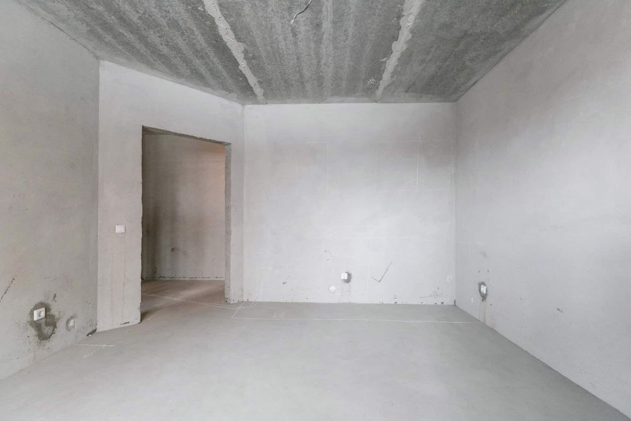 Surowe wnętrze w stanie deweloperskim: betonowy sufit, ściany przygotowane pod gładzie, widoczne instalacje elektryczne, zarysowane linie pomocnicze na podłodze, otwór drzwiowy bez ościeżnicy.