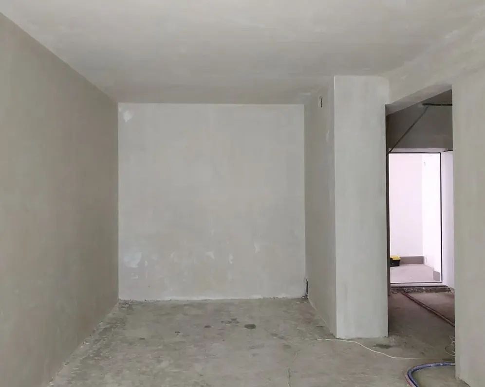Pokój w trakcie prac wykończeniowych, ściany po nałożeniu gładzi, widoczne niedoskonałości i nierówności przed malowaniem, surowa podłoga, w tle otwarte przejście do innego pomieszczenia.