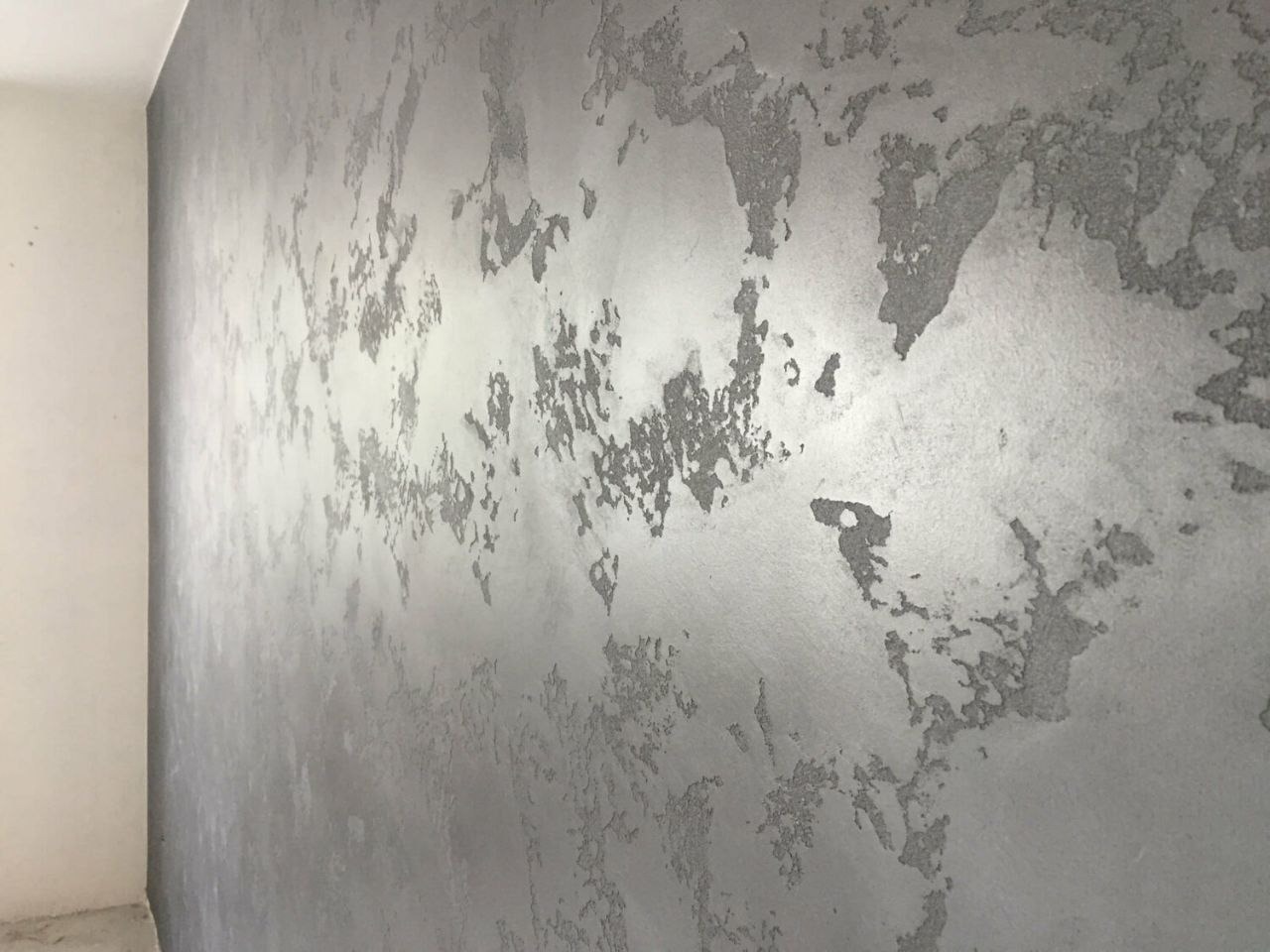 Ściana pokryta dekoracyjną, srebrzystą gładzią z wyraźną fakturą i nieregularnymi wzorami, uchwycona pod kątem, z widocznym fragmentem narożnika.