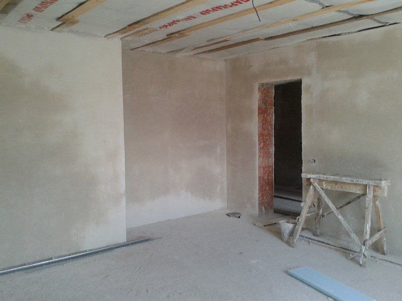 Pokój w trakcie wygładzania ścian, widoczne surowe wykończenie, drewniane belki sufitowe z folią, ściany pokryte gładzią, drewniany kozioł malarski i fragment niedokończonego przejścia z ceglaną...