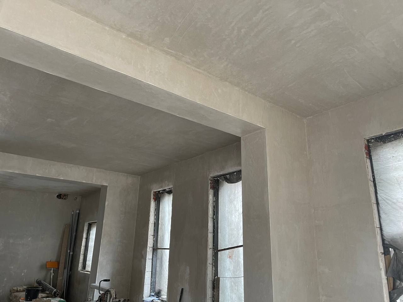 Wnętrze w trakcie prac wykończeniowych, ściany i sufit pokryte świeżo nałożoną gładzią, widoczne okna w trakcie zabezpieczania folią, na podłodze narzędzia i materiały budowlane.