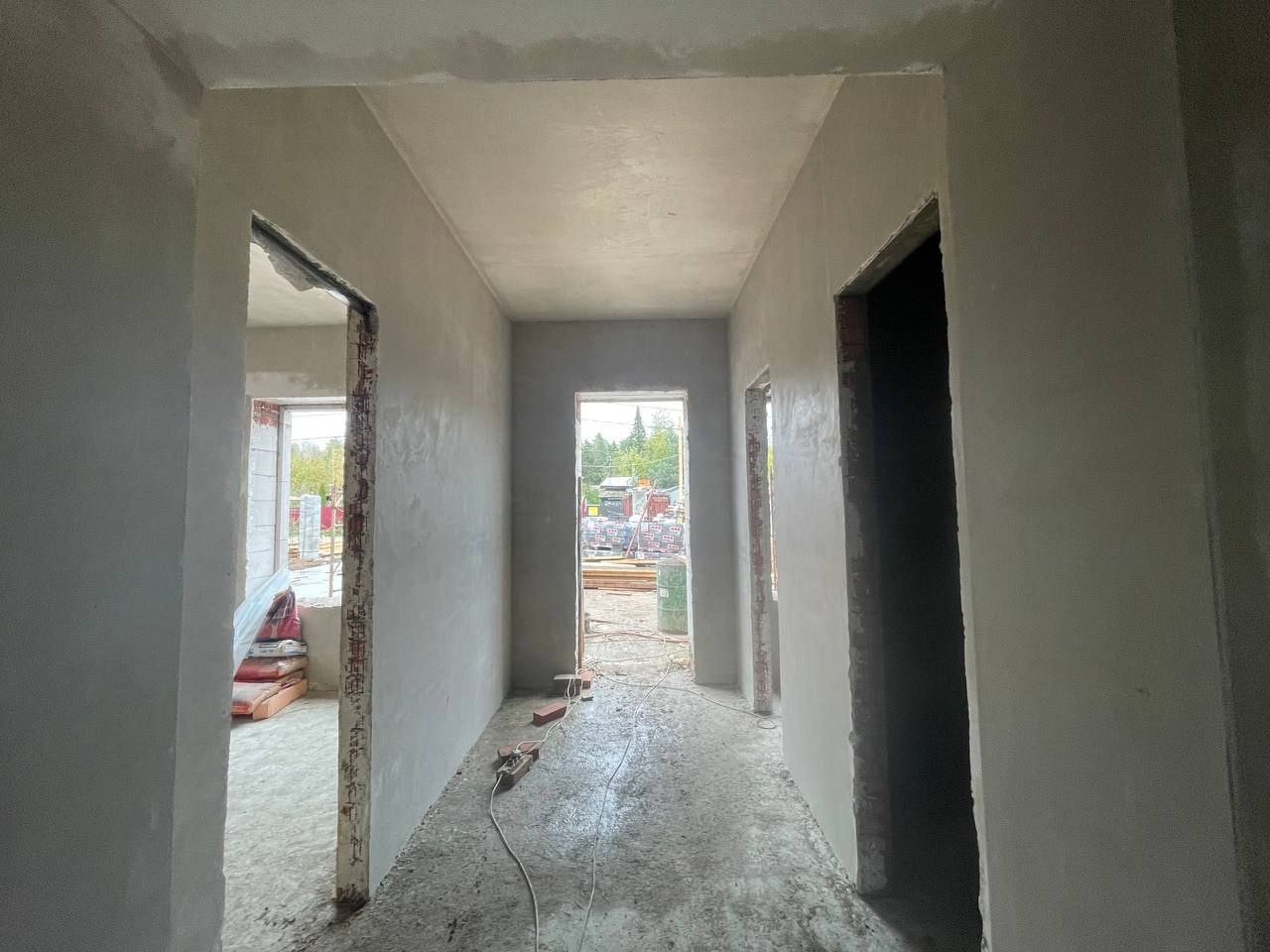 Korytarz w stanie surowym po nałożeniu gładzi, widoczne niedokończone ościeżnice drzwiowe, przewody elektryczne na podłodze i materiały budowlane w tle.