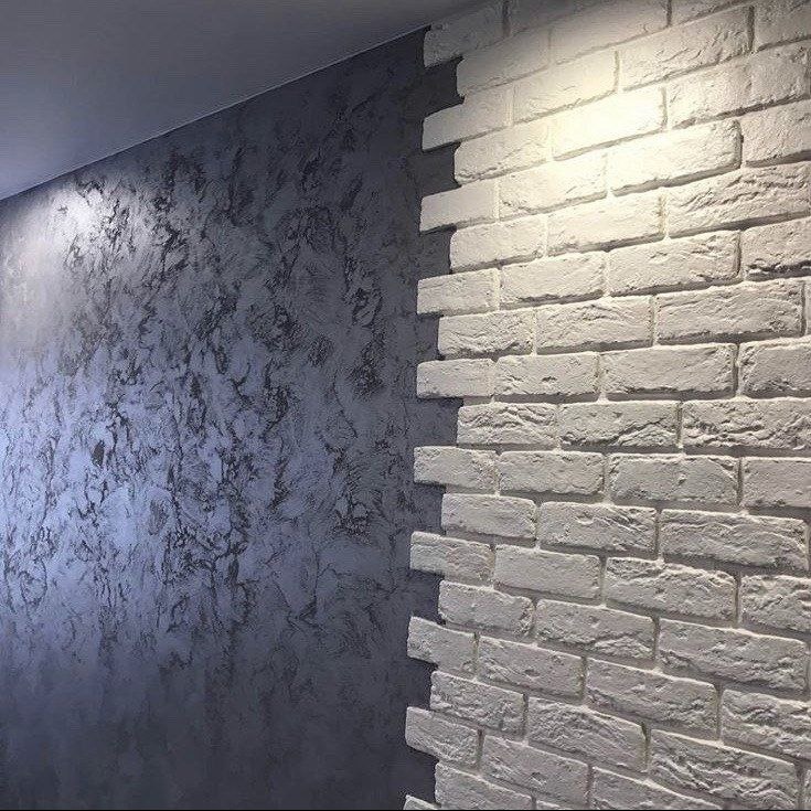 Ściana wewnętrzna z dekoracyjną gładzią w odcieniach szarości i fragmentem ściany wykończonej białymi, dekoracyjnymi płytkami imitującymi cegłę, oświetlona punktowym światłem.