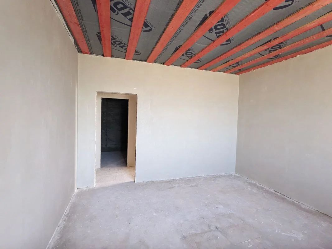 Wnętrze pokoju w stanie surowym z nałożonymi gładziami na ścianach, widoczne drewniane belki stropowe z folią izolacyjną i otwór drzwiowy bez drzwi.