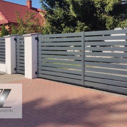 Szczygielski-Ogrodzenia - Nowoczesne, szare ogrodzenie panelowe z poziomymi przęsłami i bramą wjazdową, osadzone na białych słupkach, na tle domu z czerwoną dachówką i zielenią.