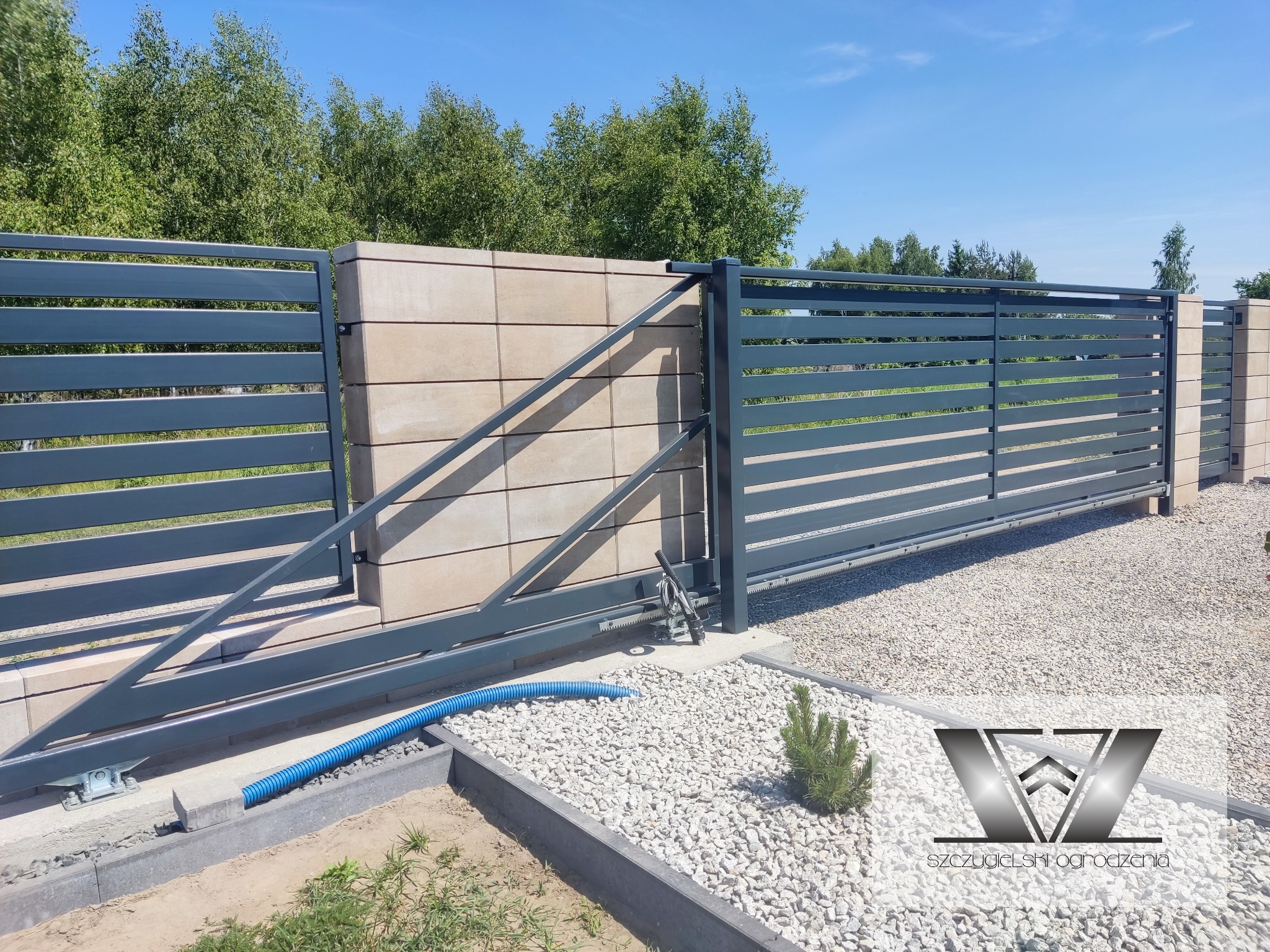 Automatyczna brama przesuwna z poziomymi panelami w kolorze grafitowym, zamontowana na słupkach z beżowych bloczków betonowych, widoczny mechanizm napędowy i niebieski przewód w peszlu.