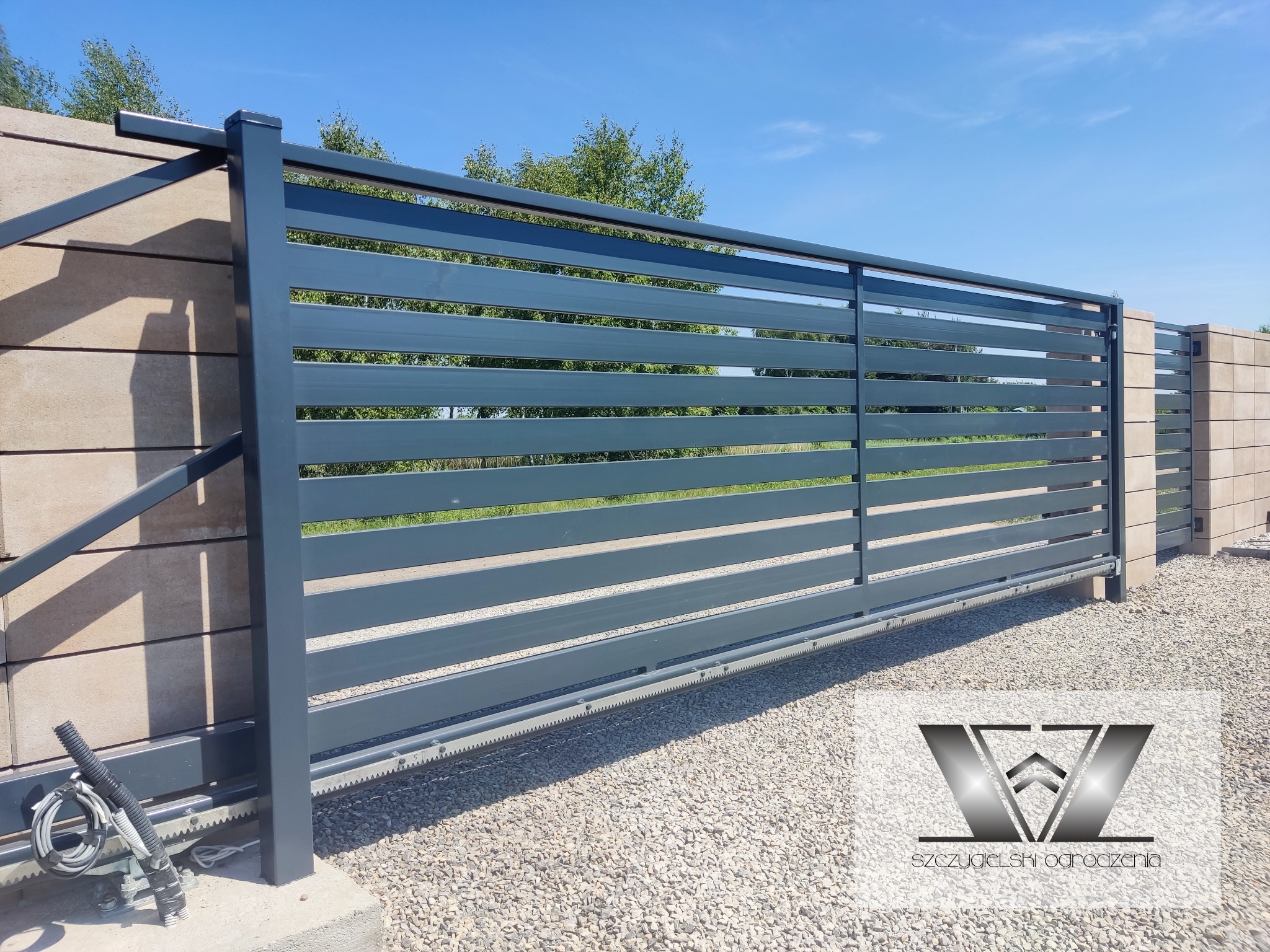 Nowoczesna, przesuwna brama w kolorze grafitowym z poziomymi panelami, zamontowana na betonowym fundamencie i prowadnicy z zębatką, z widocznym napędem elektrycznym, na tle kostki brukowej...