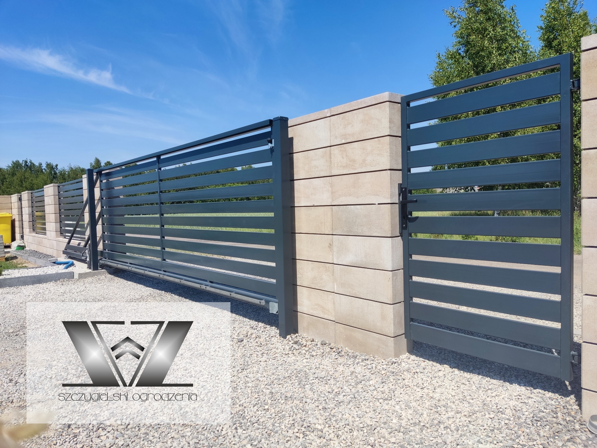 Nowoczesna brama przesuwna z poziomymi panelami w kolorze grafitowym, zamontowana między słupkami z beżowego betonu architektonicznego, widoczny mechanizm jezdny, podłoże wysypane drobnym białym...