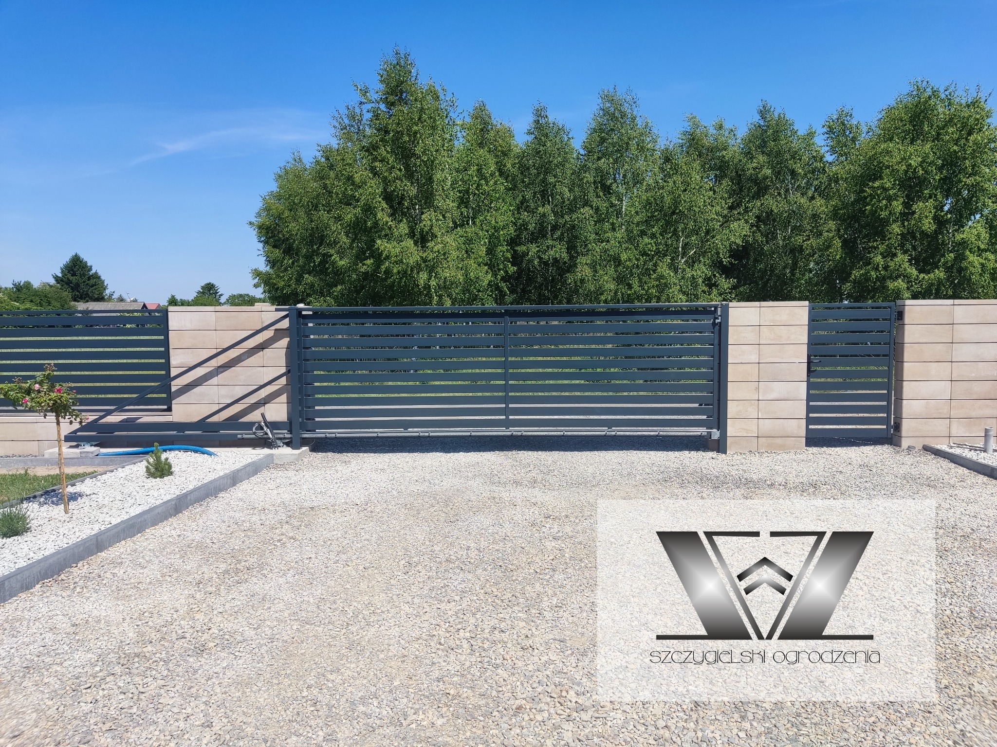 Nowoczesna, pozioma brama przesuwna z szarego metalu, osadzona w murze z beżowych bloczków betonowych, na żwirowym podłożu; w prawym dolnym rogu widoczne logo firmy.