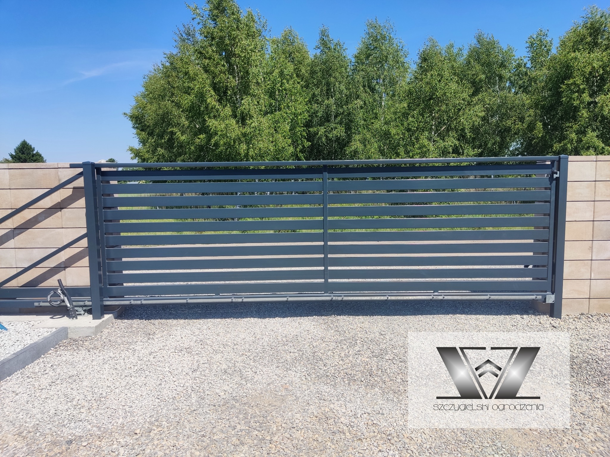 Nowoczesna, przesuwna brama wjazdowa z poziomych profili, szara, z automatyką, na tle beżowego muru i zieleni drzew.